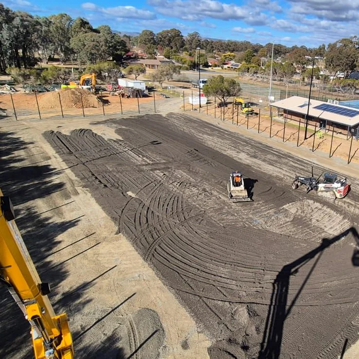 Excavator overlooking a levelled dirt site being graded by machinery before construction