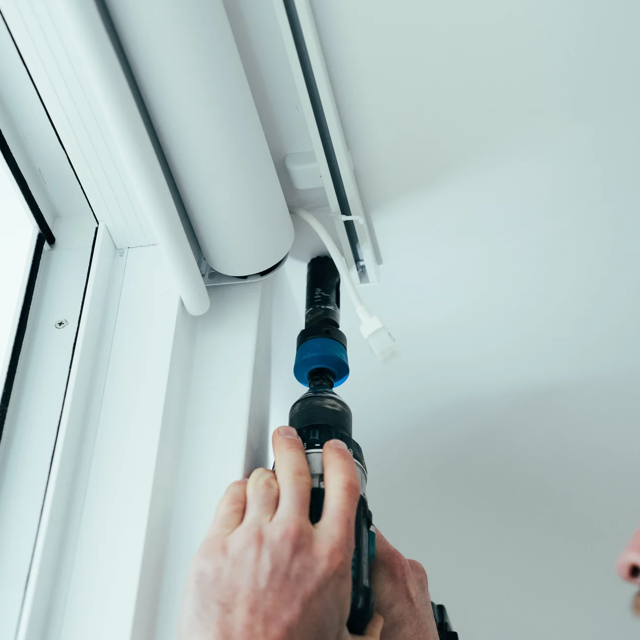 Electrician drilling into a white window frame beside a roller blind