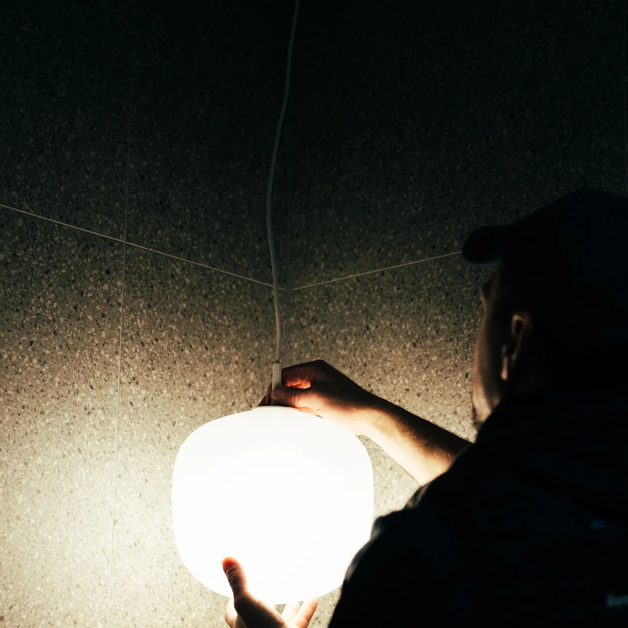 Electrician holding a glowing white globe pendant light against a speckled tiled wall