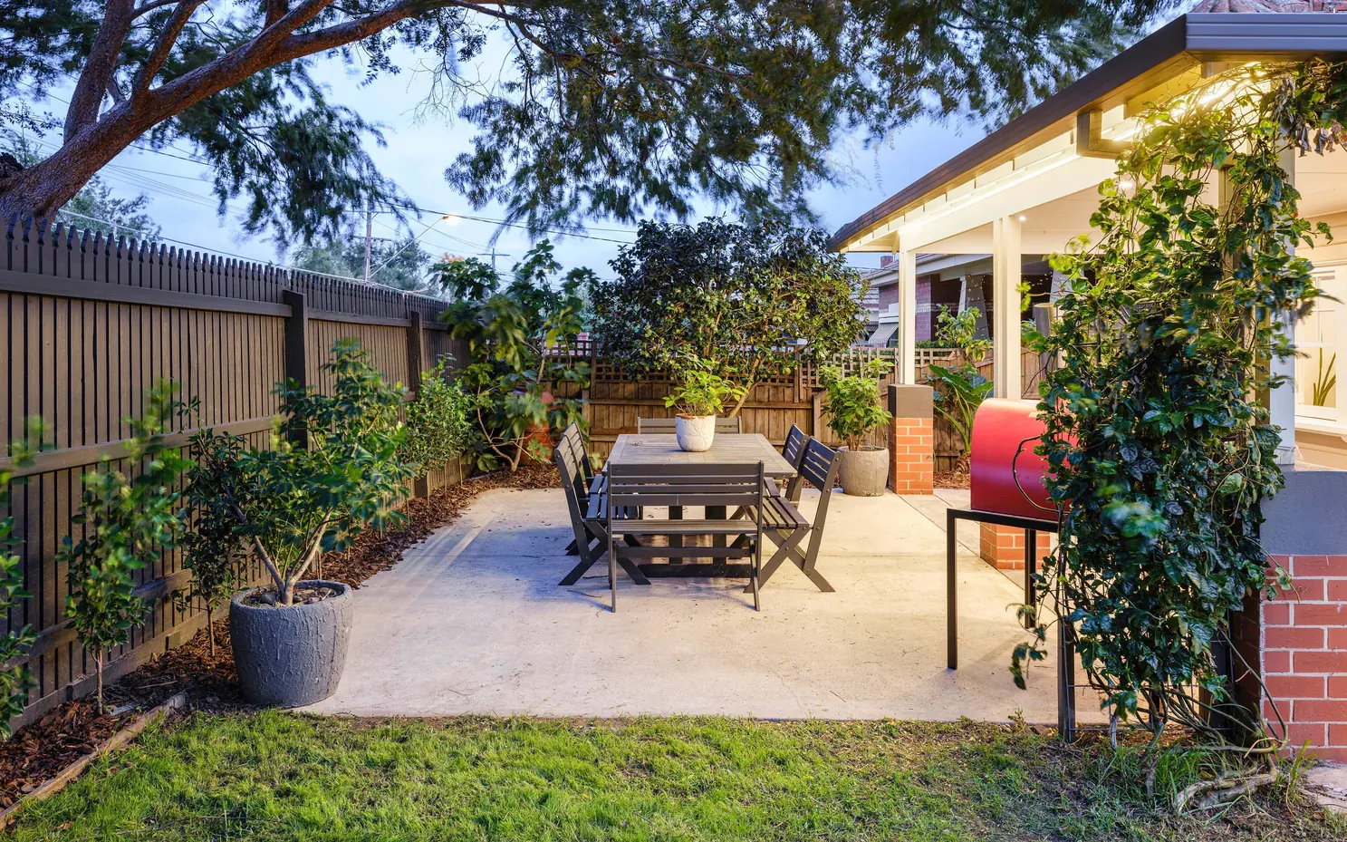 Backyard with an outdoor dining table, garden beds and a lit verandah at dusk