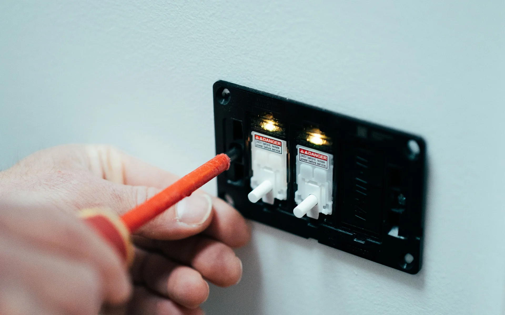 Electrician testing a black switch mechanism on a white wall with an insulated screwdriver