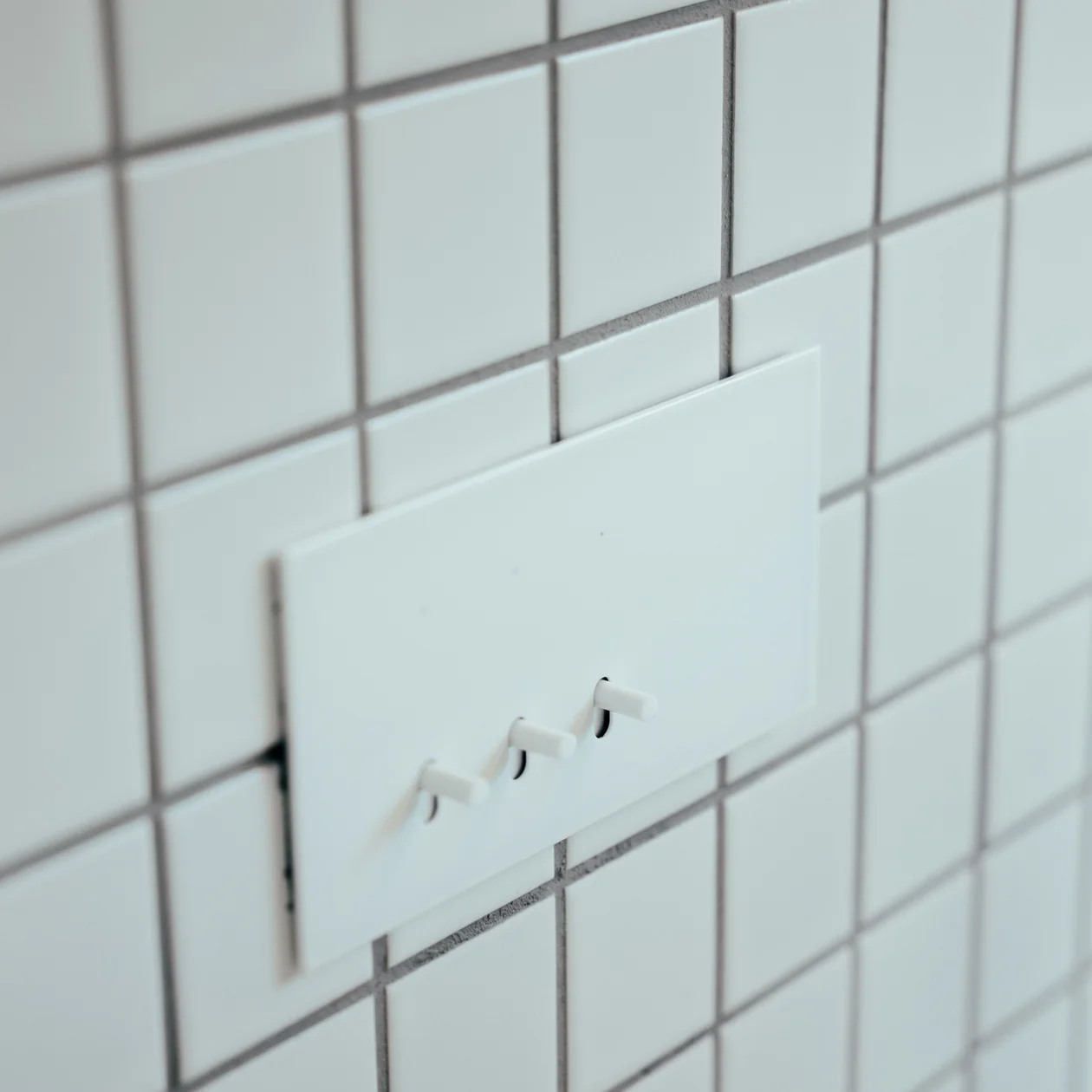 White switch plate with three toggle switches on a white square-tiled wall